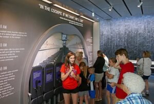 Visitors of various ages stand and observe an exhibit at the Flight 93 National Memorial visitor center, featuring a replica airplane cabin and impactful quotes from the flight, creating an immersive educational experience.