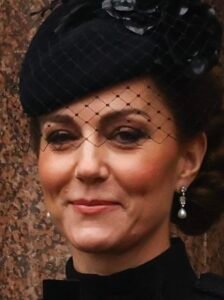 "Close-up of Catherine, Princess of Wales, in 2025, wearing a black fascinator with a mesh veil and floral detail, pearl drop earrings, and formal black attire during a public appearance."