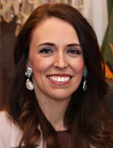 A close-up portrait of a smiling woman with long brown hair, wearing silver dangling earrings and a light-colored outfit, standing indoors with soft lighting and a blurred background.