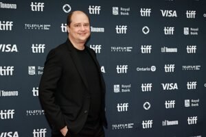 A man dressed in a black suit poses on the red carpet at the 2024 Toronto International Film Festival (TIFF), standing in front of a backdrop featuring logos of TIFF, Royal Bank, Telefilm Canada, and sponsors like VISA and Ontario Creates.