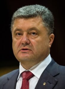 Petro Poroshenko, former President of Ukraine, speaking at a formal event in 2025. He is dressed in a dark suit with a red tie and a lapel pin of the Ukrainian coat of arms, appearing focused and serious under bright lighting.
