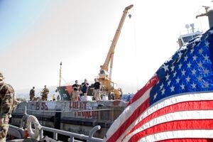 Amid rising tensions and trade disruptions, U.S. military personnel inspect port infrastructure at a harbor, symbolizing growing concern over strained supply lines. A large American flag flutters in the foreground, while a docked cargo ship and crane loom behind, underscoring the vulnerability of U.S. ports as China tariffs sharply reduce imports. The image reflects the nation’s heightened alert over potential shortages and economic impact on key maritime gateways.